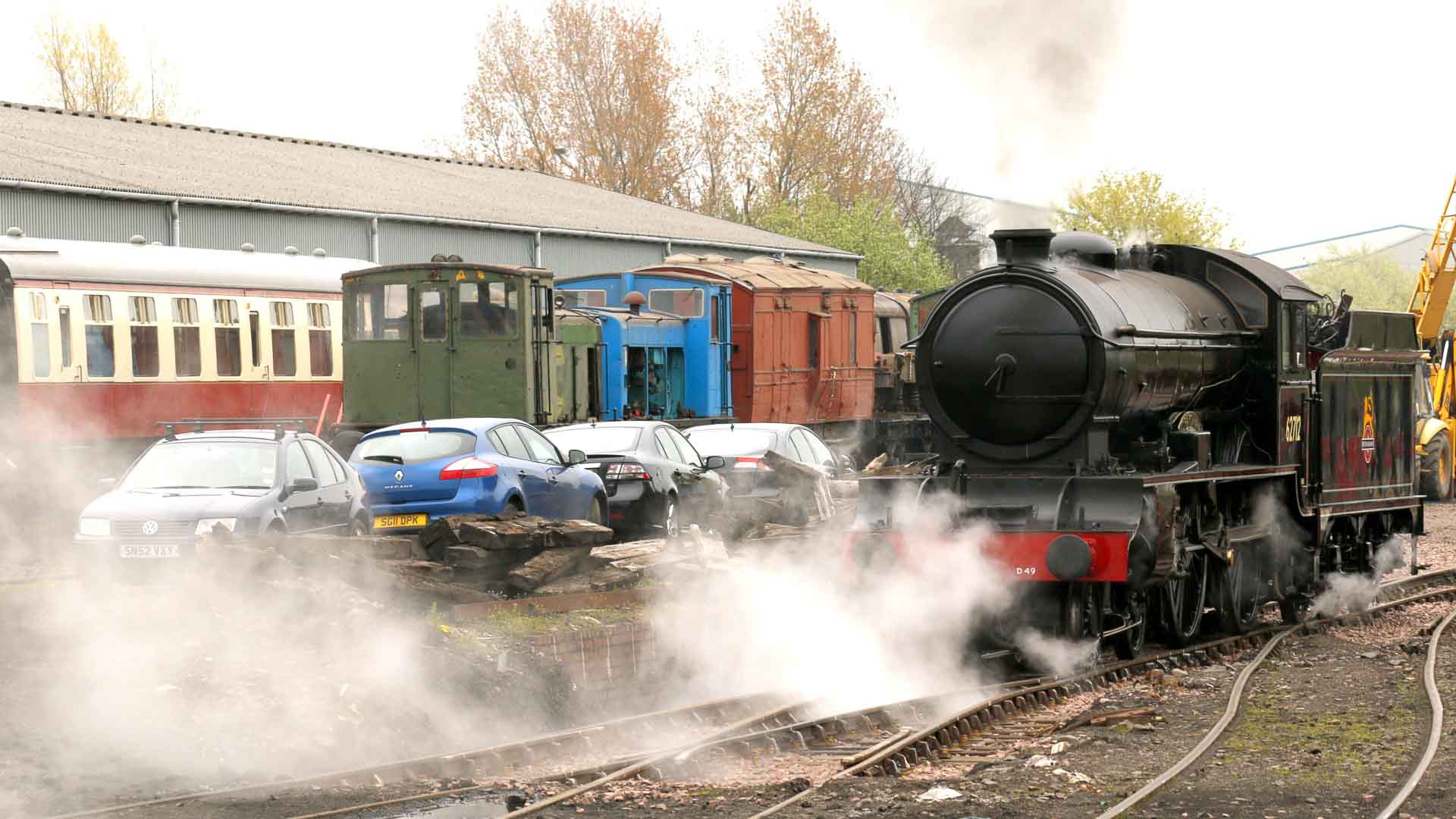 A locomotiva a vapor libera uma nuvem de vapor nos trilhos, enquanto vários carros estacionados e vagões de trem ficam ao fundo. Sob um céu nublado com árvores visíveis, evoca nostalgia em meio às tendências modernas de sustentabilidade.