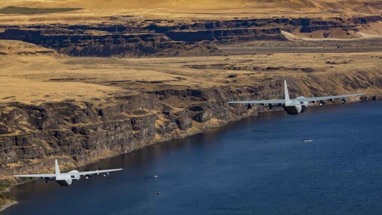 Duas aeronaves militares voam sobre um rio ladeado por penhascos, cortando a paisagem rochosa do deserto com vegetação esparsa. Sua precisão reflete a coordenação de um contêiner compartilhado bem gerenciado, navegando perfeitamente pelos desafios de importação em terrenos complexos.