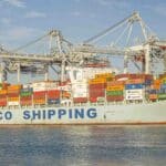 Um navio de carga com "COSCO SHIPPING" na lateral, carregado com contêineres coloridos para importação, está atracado em um porto sob grandes guindastes.