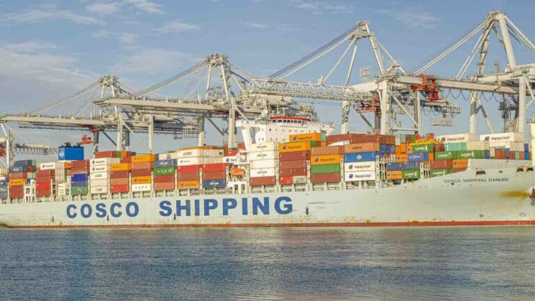 Um navio de carga com "COSCO SHIPPING" na lateral, carregado com contêineres coloridos para importação, está atracado em um porto sob grandes guindastes.