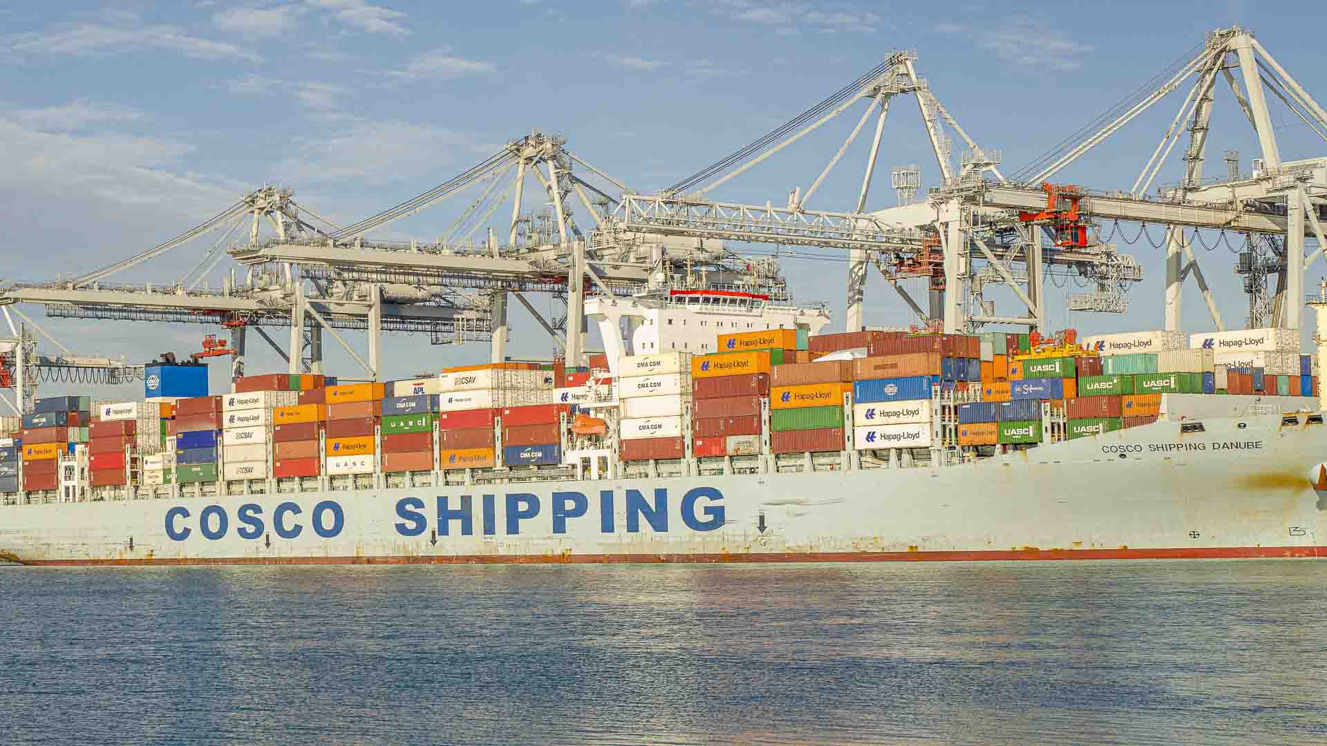 Um navio de carga com "COSCO SHIPPING" na lateral, carregado com contêineres coloridos para importação, está atracado em um porto sob grandes guindastes.