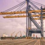 Um estaleiro com grandes guindastes elevando-se sobre contêineres, exibindo a atividade movimentada de uma empresa focada em bens importados, com um navio de carga atracado ao fundo sob um céu nublado.