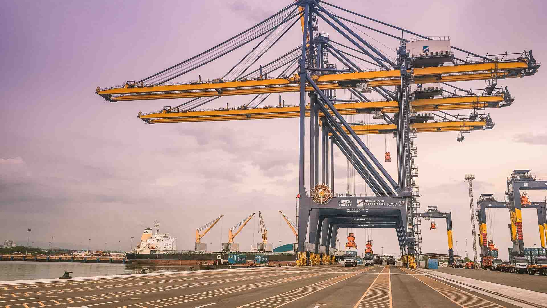 Um estaleiro com grandes guindastes elevando-se sobre contêineres, exibindo a atividade movimentada de uma empresa focada em bens importados, com um navio de carga atracado ao fundo sob um céu nublado.