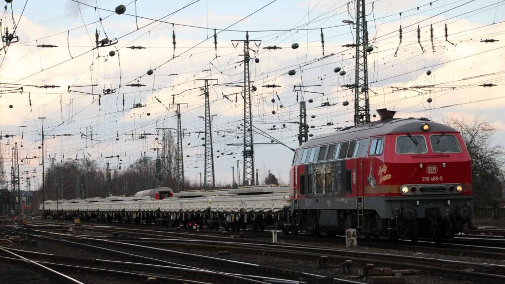 Um trem vermelho com vários vagões viaja pelos trilhos sob uma rede de fios aéreos, em um cenário com céu nublado, que lembra as movimentadas rotas usadas para importar da China.