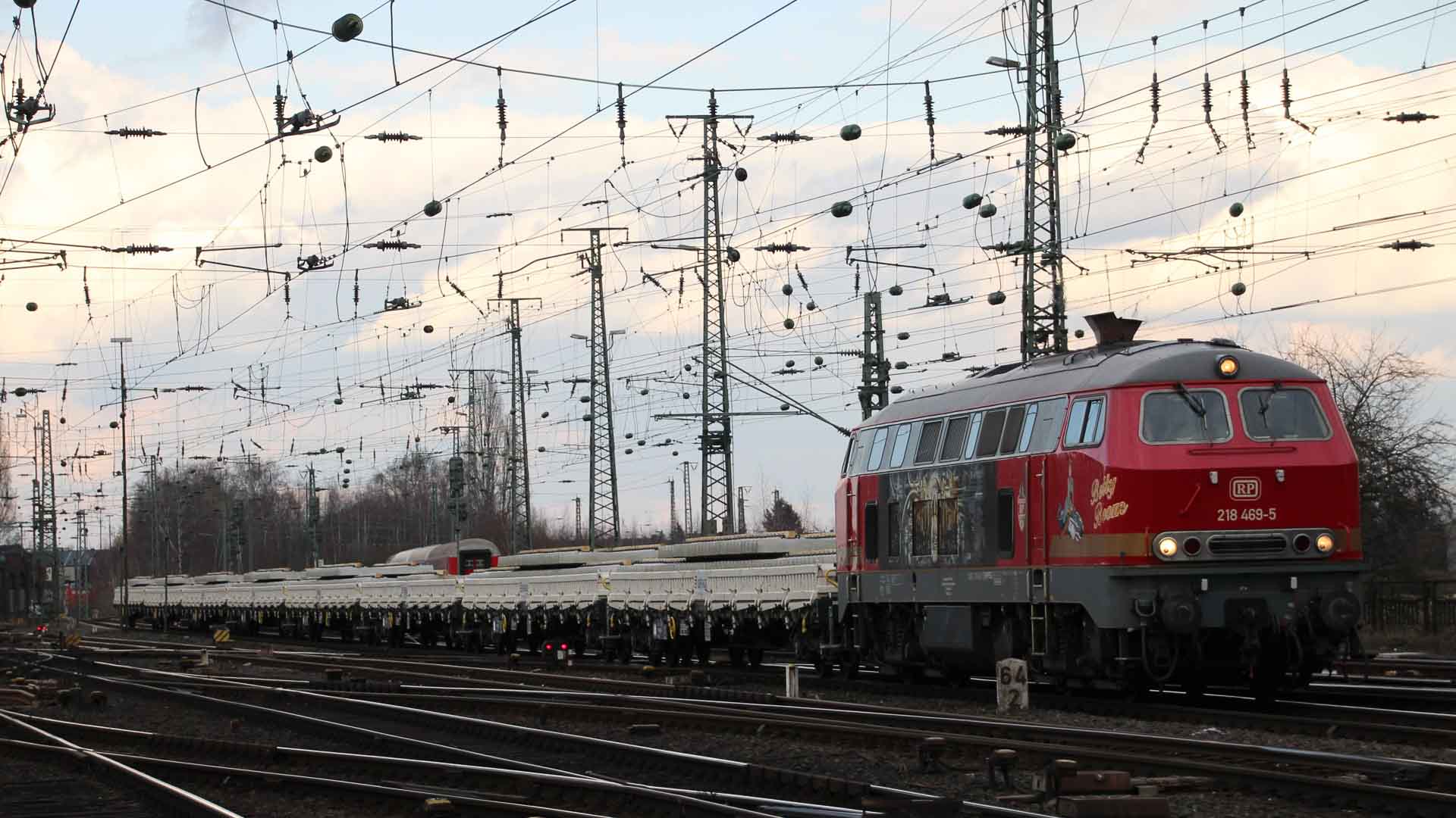Um trem vermelho com vários vagões viaja pelos trilhos sob uma rede de fios aéreos, em um cenário com céu nublado, que lembra as movimentadas rotas usadas para importar da China.