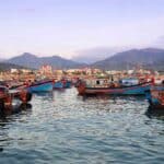 Uma cena colorida de um porto com vários barcos de pesca ancorados em águas calmas perto de um píer de madeira, com um cenário montanhoso sob um céu parcialmente nublado, que lembra cenários costeiros na China.