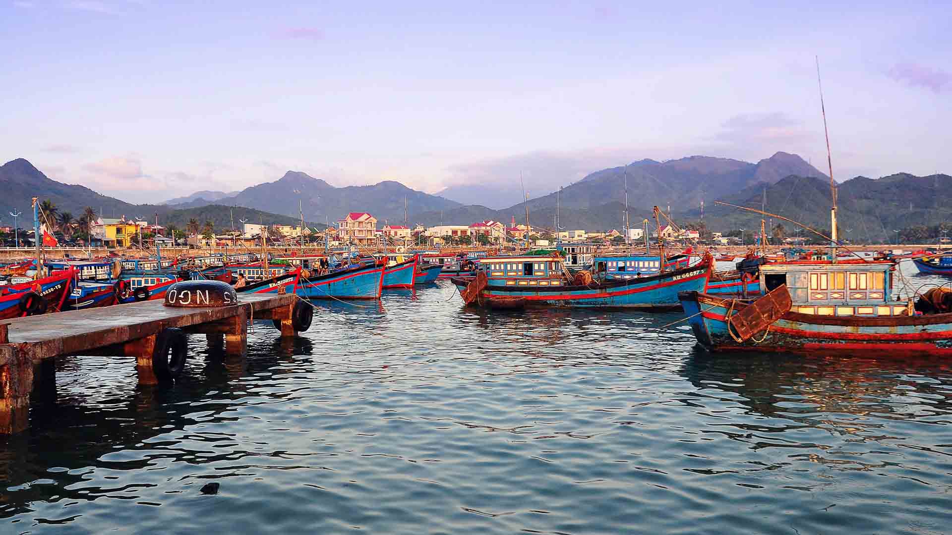 Uma cena colorida de um porto com vários barcos de pesca ancorados em águas calmas perto de um píer de madeira, com um cenário montanhoso sob um céu parcialmente nublado, que lembra cenários costeiros na China.