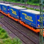 Um trem de carga com contêineres azuis e amarelos, simbolizando importação empresarial, viaja sobre trilhos por uma paisagem verdejante.