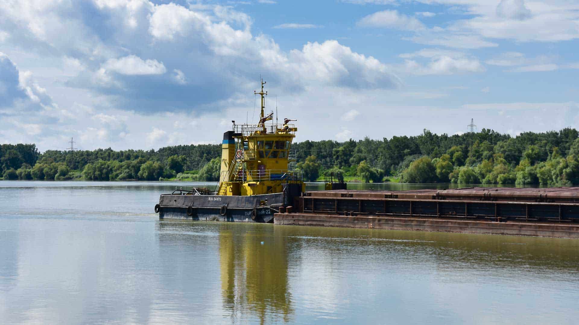 Um rebocador amarelo, crucial para a importação em 2024, empurra uma grande barcaça em um rio calmo, cercado por árvores e sob um céu parcialmente nublado.