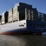 Um grande navio de carga carregado com contêineres de transporte empilhados navega calmamente em um rio sob um céu limpo. Prédios distantes alinham-se na margem, permanecendo como lembretes do mundo próspero dos importadores e das lições essenciais aprendidas no comércio global.