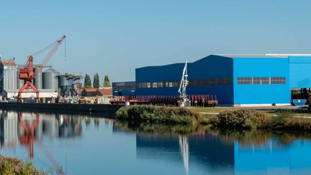 Uma cena industrial à beira-mar com um armazém azul, um guindaste vermelho e vários silos pinta o quadro enquanto olhamos para 2025. Águas calmas refletem essas estruturas, sugerindo práticas de importação refinadas sob um céu azul claro ao fundo.