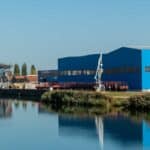 Uma cena industrial à beira-mar com um armazém azul, um guindaste vermelho e vários silos pinta o quadro enquanto olhamos para 2025. Águas calmas refletem essas estruturas, sugerindo práticas de importação refinadas sob um céu azul claro ao fundo.