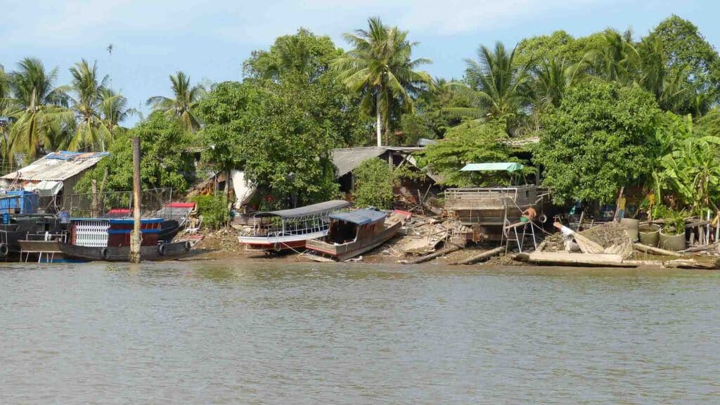 Barcos atracados ao longo de uma margem lamacenta de rio cercada por vegetação e árvores exuberantes, com estruturas de madeira e folhagens ao fundo sob um céu claro, criam um cenário idílico perfeito para considerar quais são os melhores produtos para importar e revender.