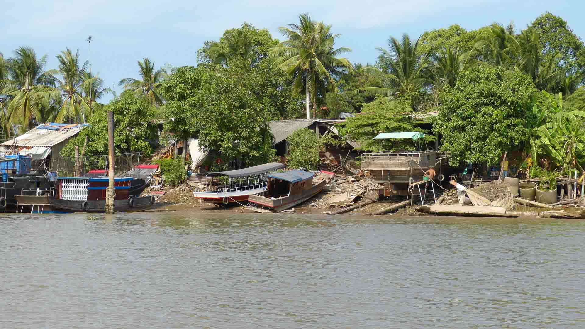 Barcos atracados ao longo de uma margem lamacenta de rio cercada por vegetação e árvores exuberantes, com estruturas de madeira e folhagens ao fundo sob um céu claro, criam um cenário idílico perfeito para considerar quais são os melhores produtos para importar e revender.