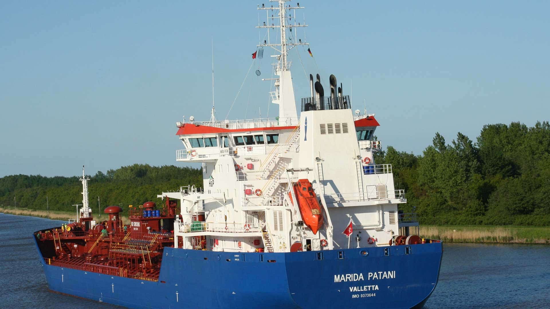 O navio de carga azul e vermelho chamado "Marida Patani" desliza pacificamente em um canal calmo, com árvores verdes ao longo de seu caminho. Conforme o navio embarca em sua viagem, as compras internacionais a bordo sugerem como o comércio global pode em breve se ajustar às políticas da Nova Taxação.