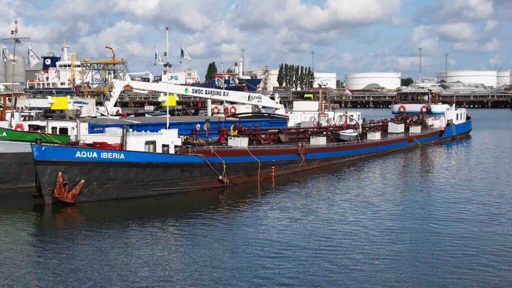 Uma barcaça chamada Aqua Iberia atracou em um porto industrial, cercada por outras embarcações, causando um impacto significativo no espaço movimentado, com grandes tanques de armazenamento se destacando ao fundo.