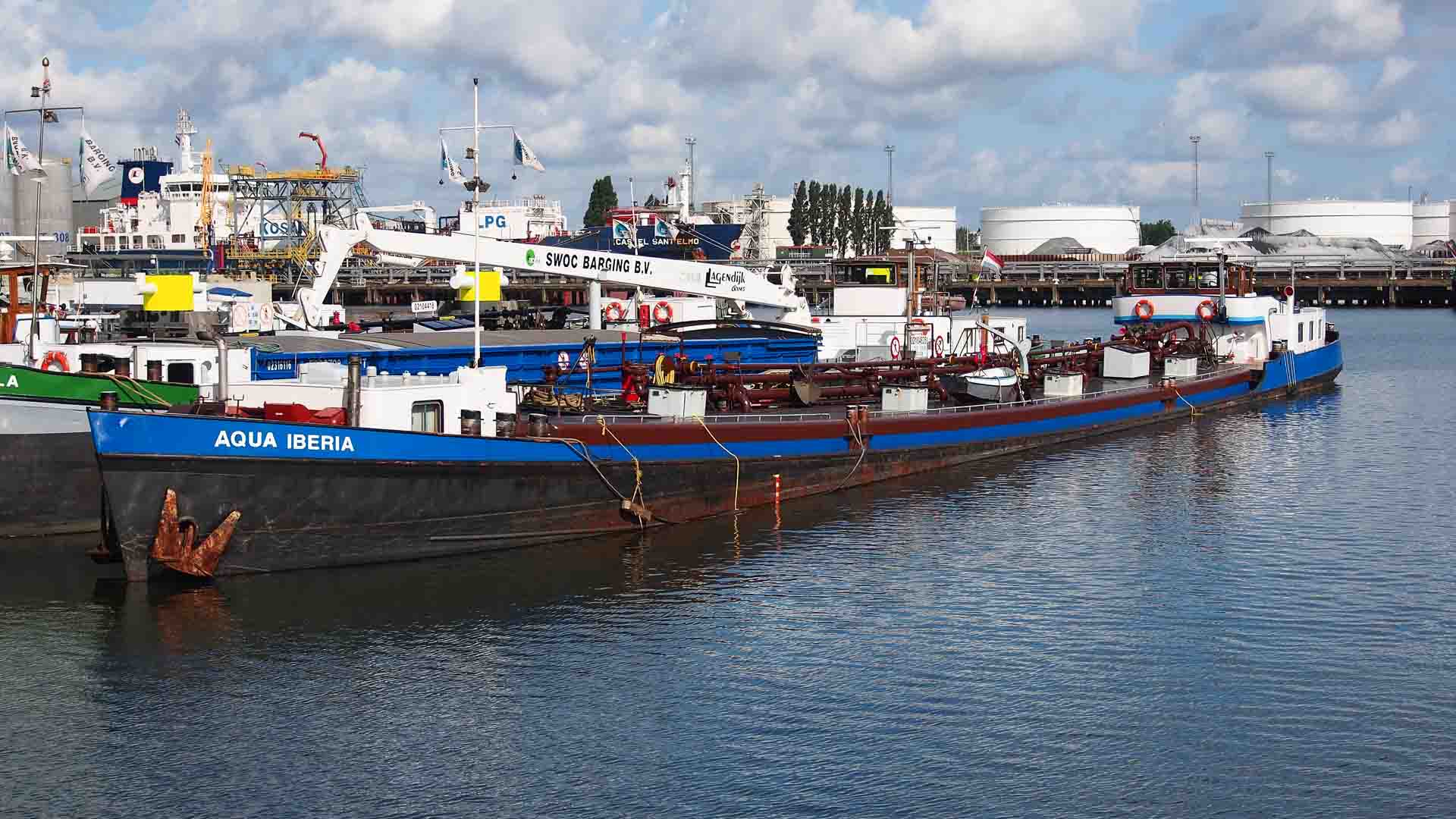 Uma barcaça chamada Aqua Iberia atracou em um porto industrial, cercada por outras embarcações, causando um impacto significativo no espaço movimentado, com grandes tanques de armazenamento se destacando ao fundo.