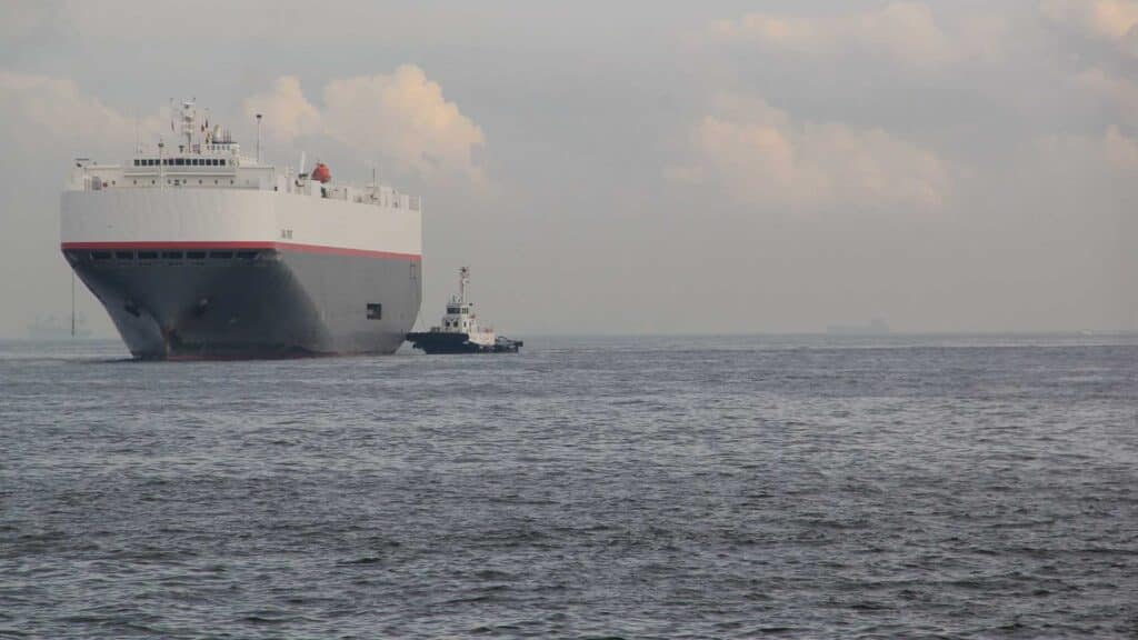 Um grande navio de carga, essencial para importar mercadorias da China, navega ao lado de um pequeno rebocador em um mar calmo sob um céu nublado.