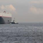 Um grande navio de carga, essencial para importar mercadorias da China, navega ao lado de um pequeno rebocador em um mar calmo sob um céu nublado.