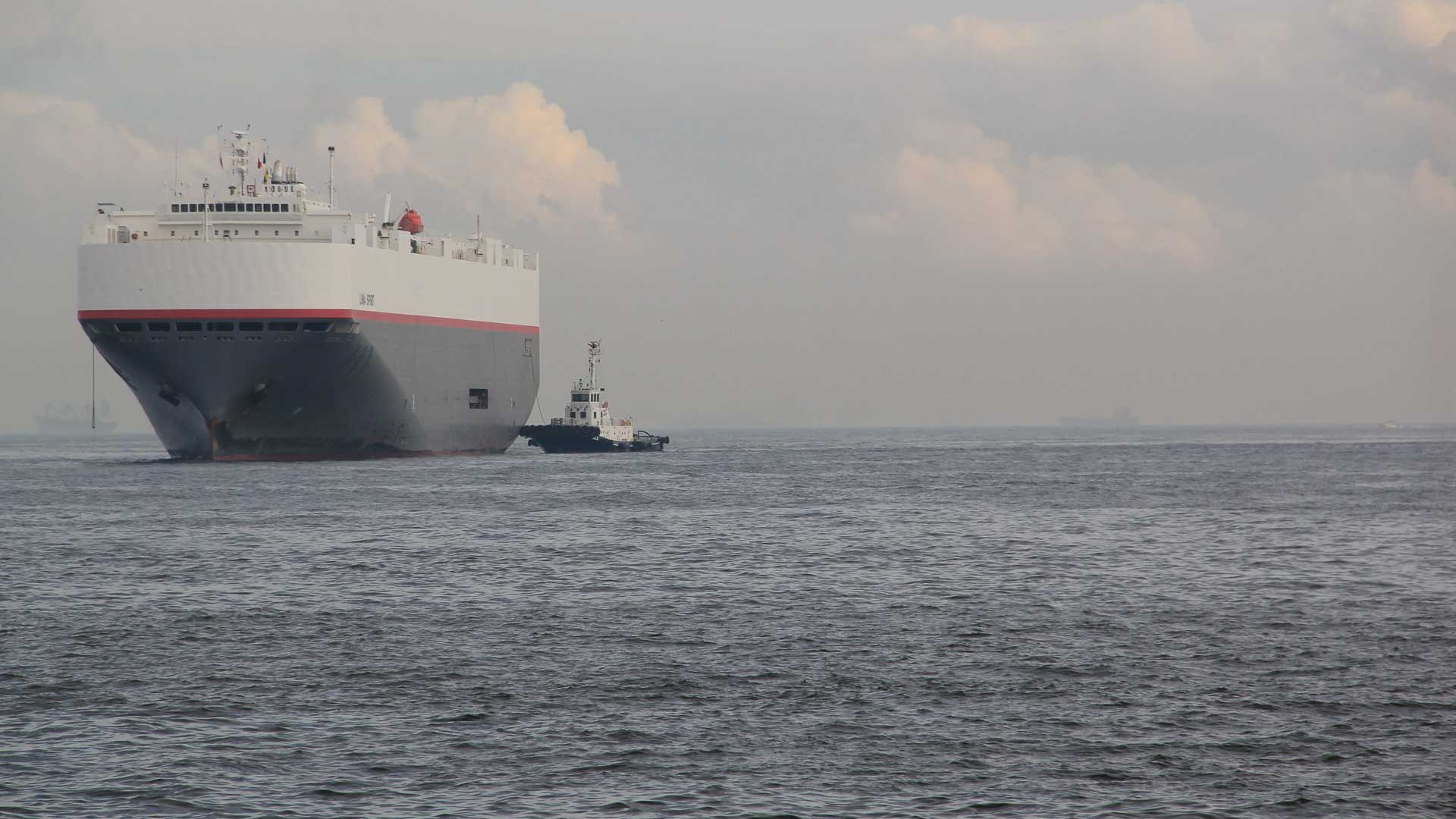 Um grande navio de carga, essencial para importar mercadorias da China, navega ao lado de um pequeno rebocador em um mar calmo sob um céu nublado.
