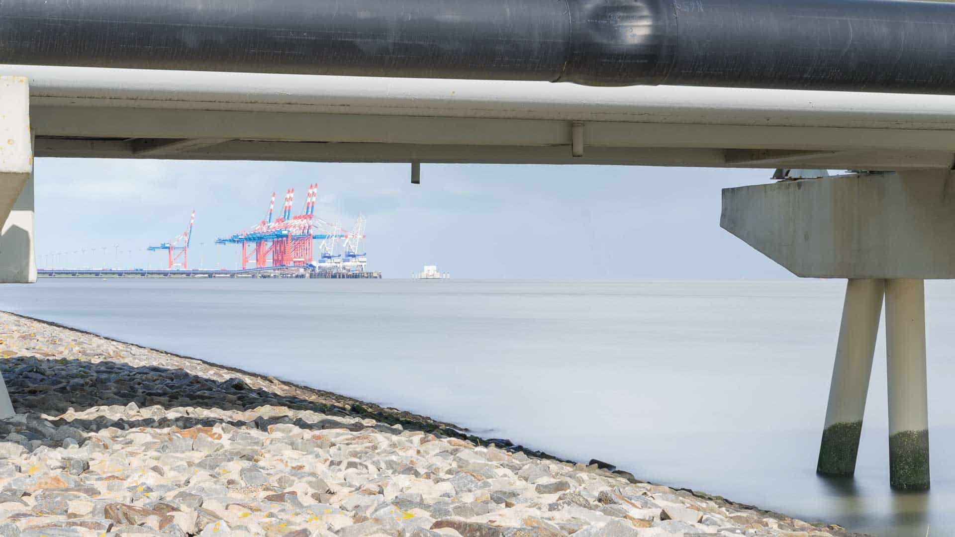 Sob a ponte, a costa rochosa se estende com distantes guindastes industriais visíveis contra um céu nublado, destacando a importância dos produtos que abastecem o movimentado porto.