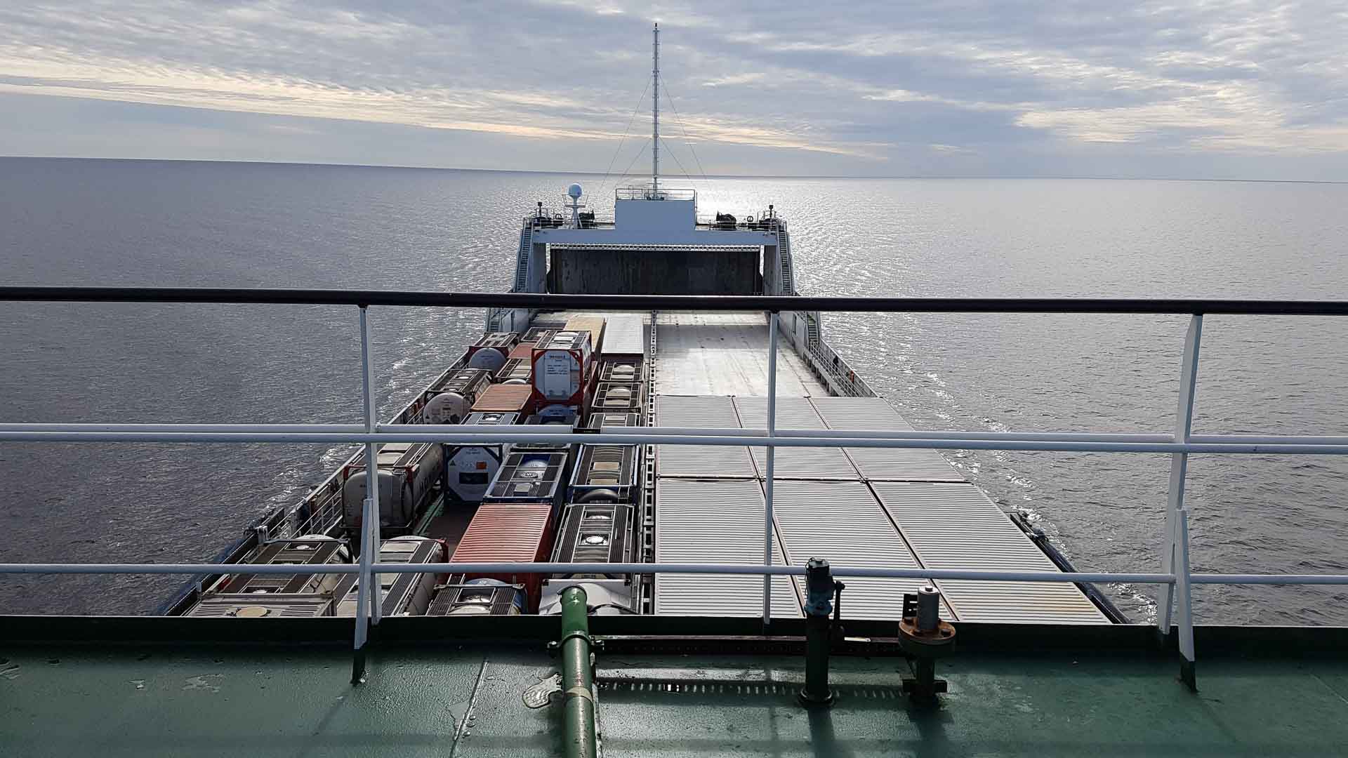 Vista do convés de um navio com vista para o mar, com contêineres de transporte empilhados a bordo, necessários para o comércio global. A água calma sob um céu nublado pinta uma cena tranquila, nos lembrando do papel vital que os navios desempenham na importação de bens em todo o mundo.