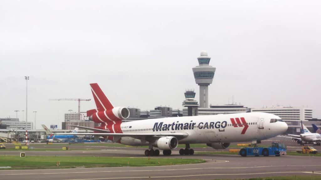 Um avião com a pintura "Martinair Cargo" está na pista de um aeroporto, pronto para sua viagem à China, com a torre de controle e outros edifícios do aeroporto visíveis ao fundo.