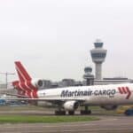 Um avião com a pintura "Martinair Cargo" está na pista de um aeroporto, pronto para sua viagem à China, com a torre de controle e outros edifícios do aeroporto visíveis ao fundo.