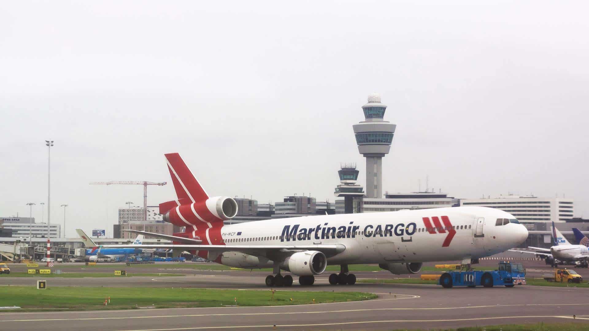 Um avião com a pintura "Martinair Cargo" está na pista de um aeroporto, pronto para sua viagem à China, com a torre de controle e outros edifícios do aeroporto visíveis ao fundo.