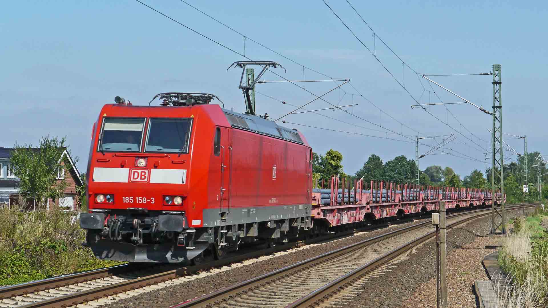 Um trem de carga elétrico vermelho, movido a energia limpa, viaja sobre trilhos por uma paisagem rural, destacando o verde intenso das árvores ao fundo.