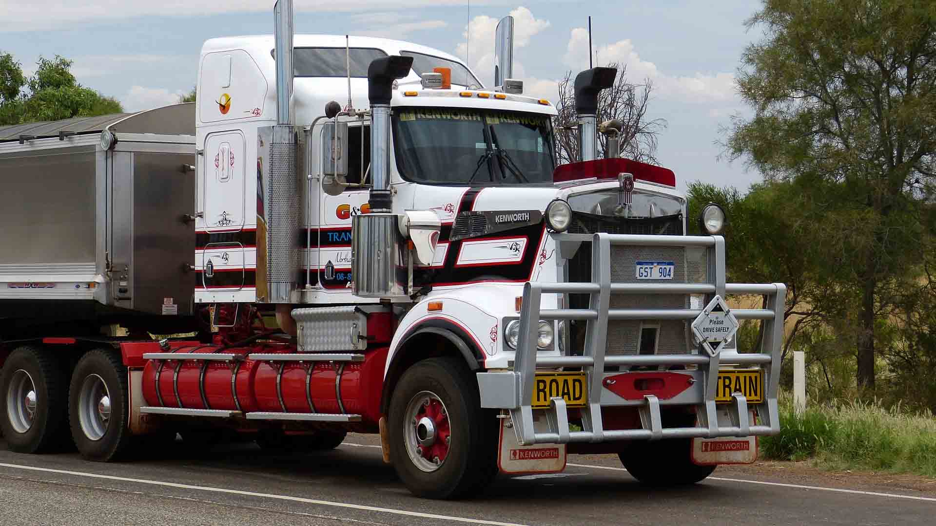 Um grande caminhão vermelho e branco com a etiqueta "Road Train", posicionado como um símbolo de importação, estacionado na beira da estrada com árvores ao fundo.