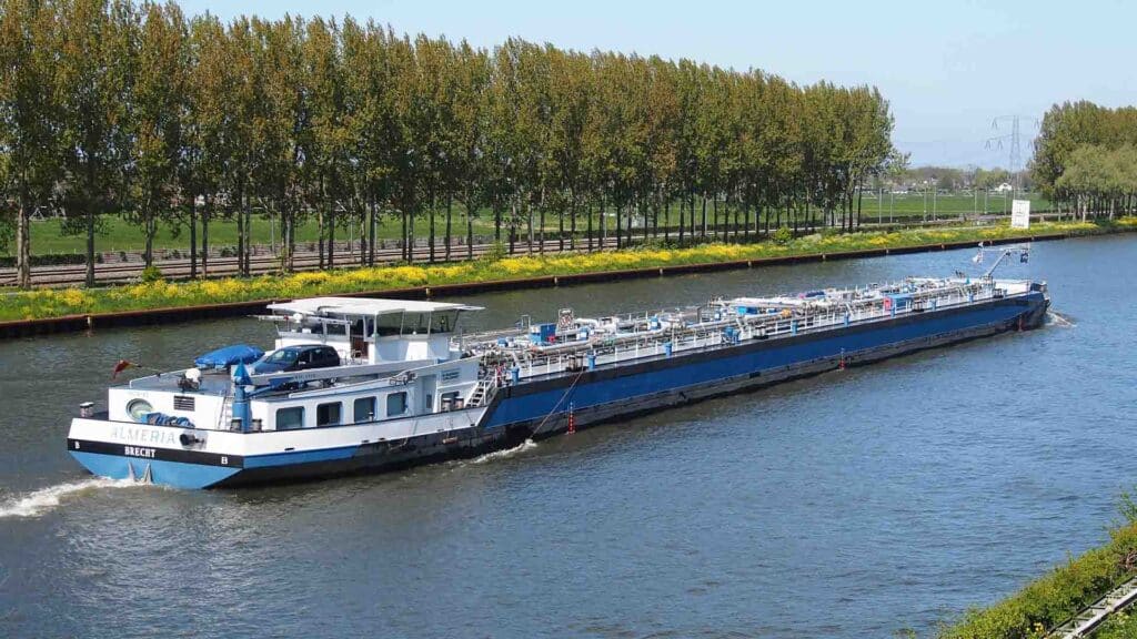 Uma grande barcaça de carga azul e branca, transportando produtos importados, viaja ao longo de um amplo canal ladeado por árvores verdes e uma paisagem gramada sob um céu claro.