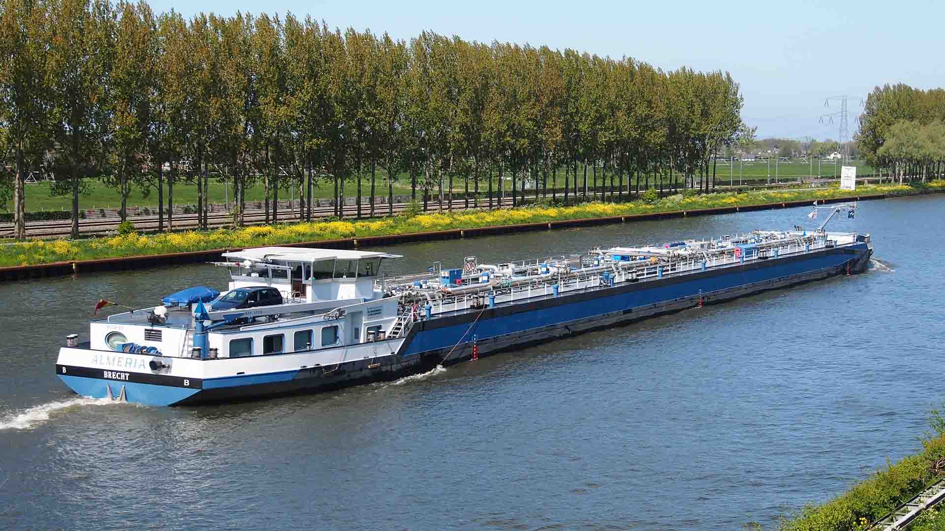 Uma grande barcaça de carga azul e branca, transportando produtos importados, viaja ao longo de um amplo canal ladeado por árvores verdes e uma paisagem gramada sob um céu claro.