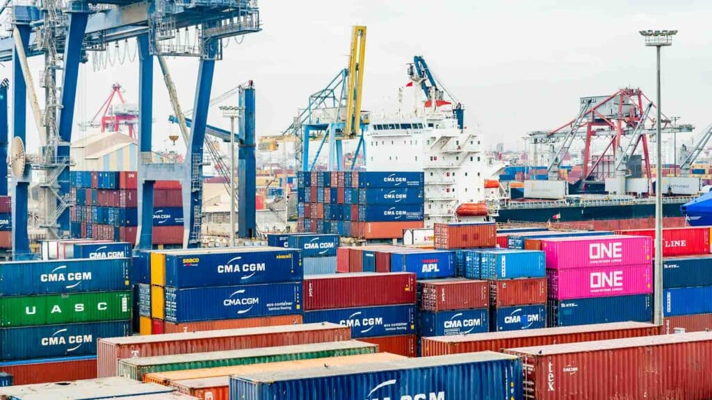 Um movimentado porto de embarque com vários contêineres de carga empilhados em várias cores aguarda aqueles que procuram visitar fornecedores chineses. Grandes guindastes dominam a cena, e um navio está atracado ao fundo, pronto para se conectar com fornecedores globais.