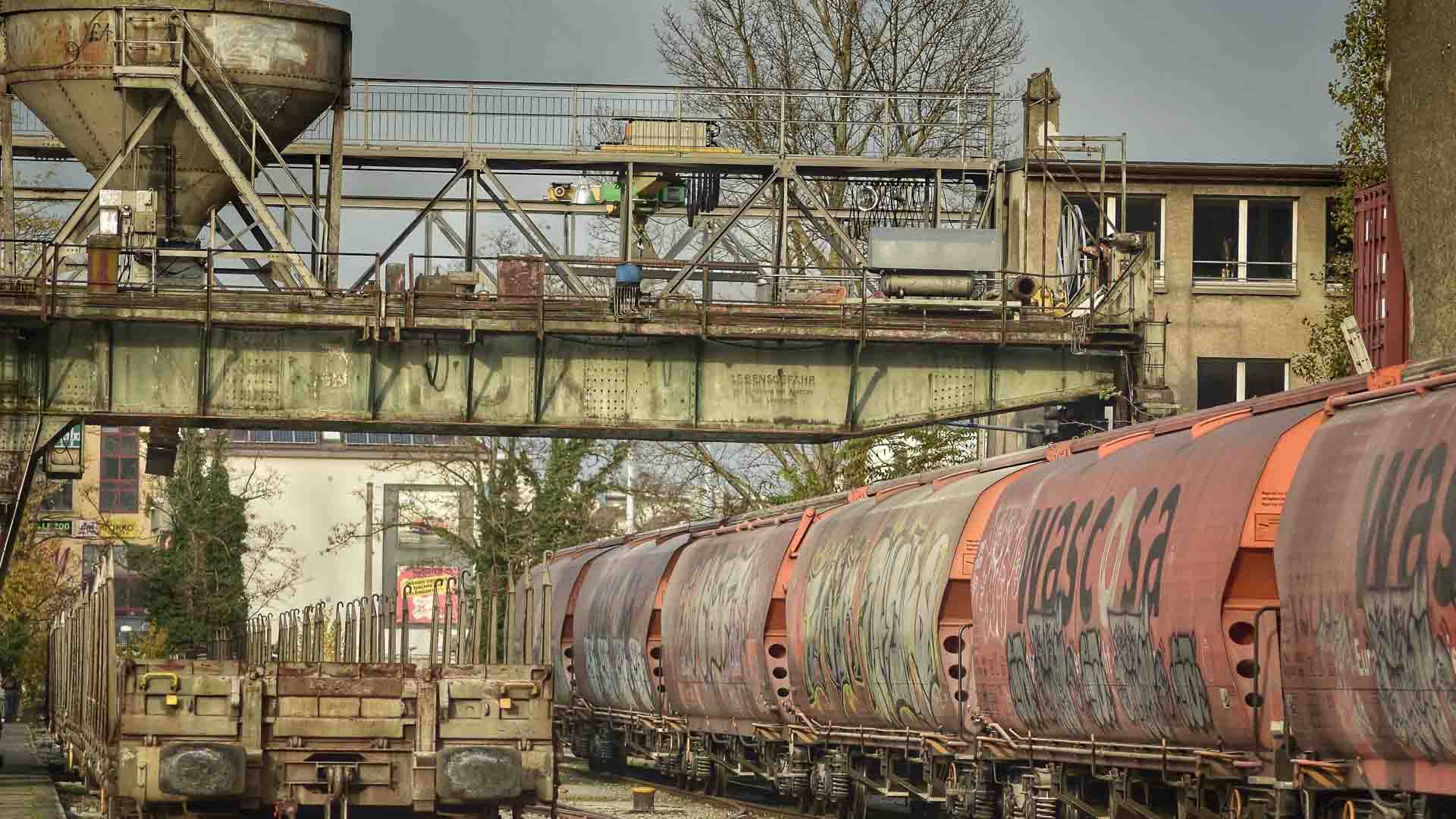 Vagões de trem de carga cobertos de pichações passam sob uma ponte industrial enferrujada em um ambiente urbano, com prédios e árvores ao fundo — uma cena que lembra os movimentados mercados chineses ou feiras na China em meio a um cenário de fornecedores diversos.