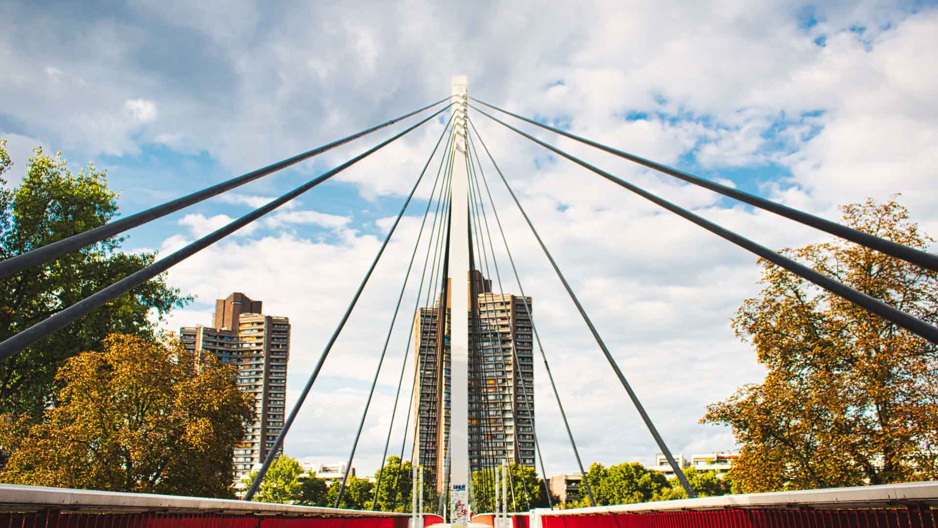 Uma estrutura geométrica de ponte com cabos, ladeada por árvores, leva a edifícios altos sob um céu parcialmente nublado, lembrando o design eficiente frequentemente encontrado em produtos chineses acessíveis.