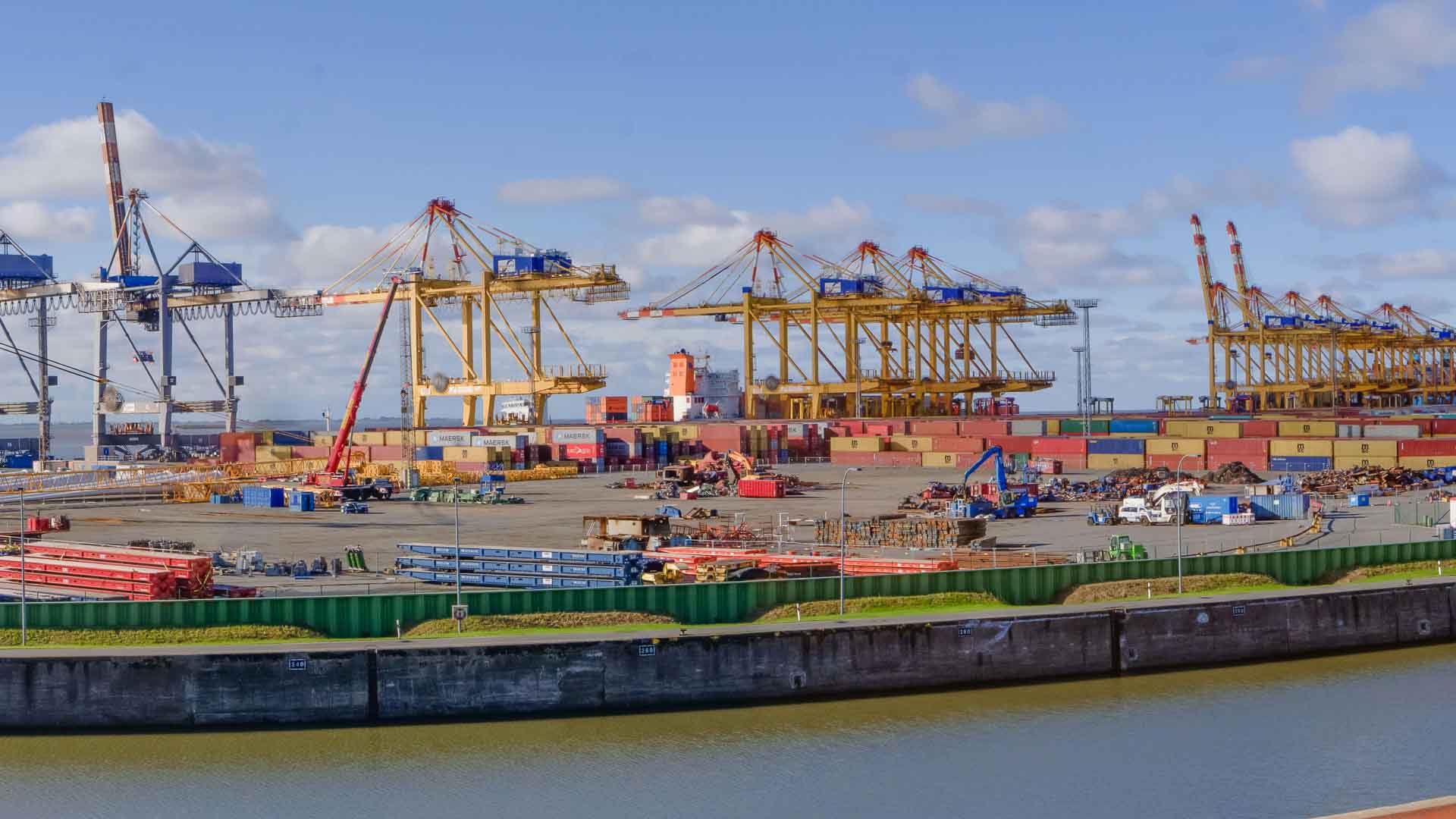 Uma movimentada cena portuária com guindastes imponentes, Produtos da China empilhados no alto em contêineres coloridos e equipamentos industriais sob um céu azul claro. A hidrovia brilha em primeiro plano, enquanto construções e veículos aumentam a atividade em toda a área.