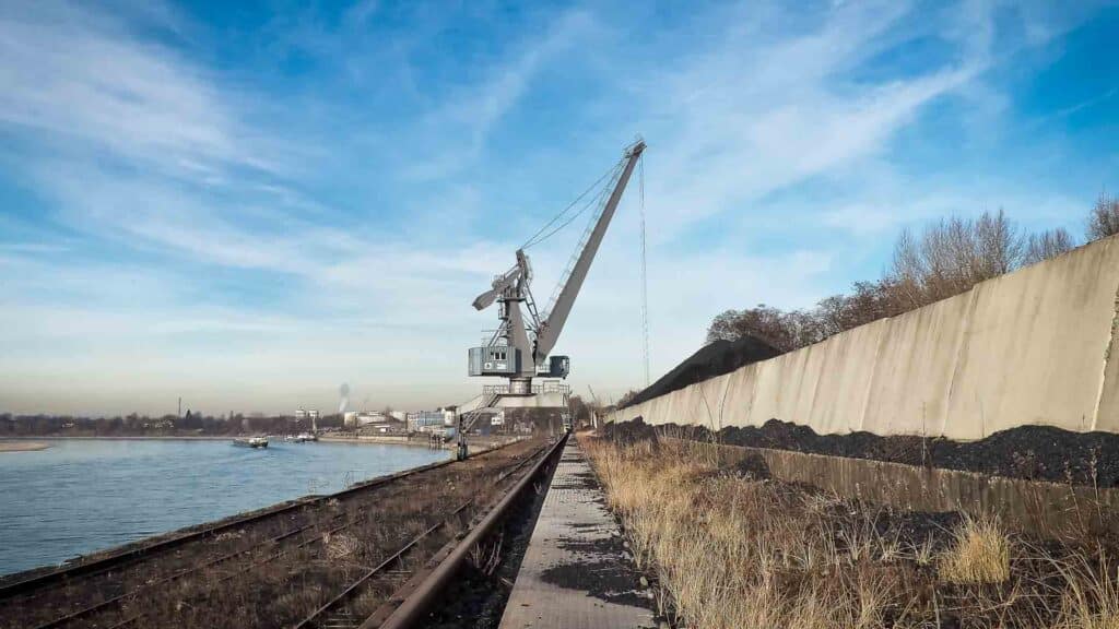 Um grande guindaste fica ao lado de um rio e de um caminho de cascalho com trilhos, lembrando as cenas movimentadas de 25 de março. Pilhas de material estão empilhadas ao longo de uma parede de concreto sob um céu azul com nuvens, competindo por atenção contra o vibrante cenário.
