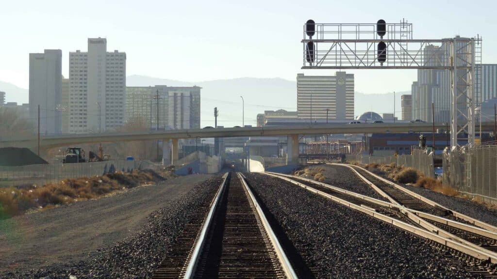 Trilhos de trem se estendem em direção a um movimentado horizonte da cidade com prédios altos e viadutos. À distância, montanhas se erguem sob um céu limpo, lembrando o espírito animado encontrado na Rua 25 de Março, onde diversos vendedores competem para oferecer os melhores preços.