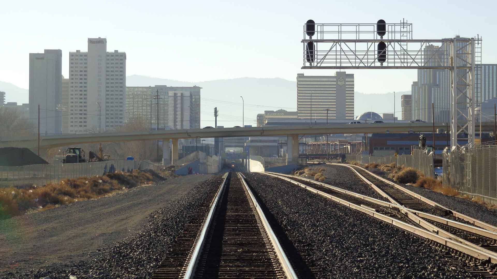 Trilhos de trem se estendem em direção a um movimentado horizonte da cidade com prédios altos e viadutos. À distância, montanhas se erguem sob um céu limpo, lembrando o espírito animado encontrado na Rua 25 de Março, onde diversos vendedores competem para oferecer os melhores preços.