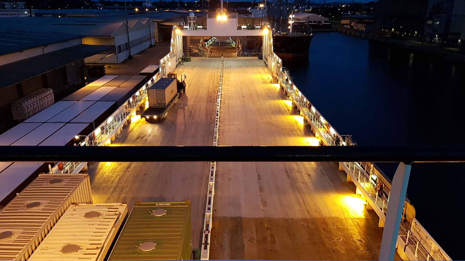 O convés de um navio de carga, transportando produtos importados, é iluminado à noite, com veículos e contêineres visíveis. Uma empilhadeira navega no lado esquerdo do convés.