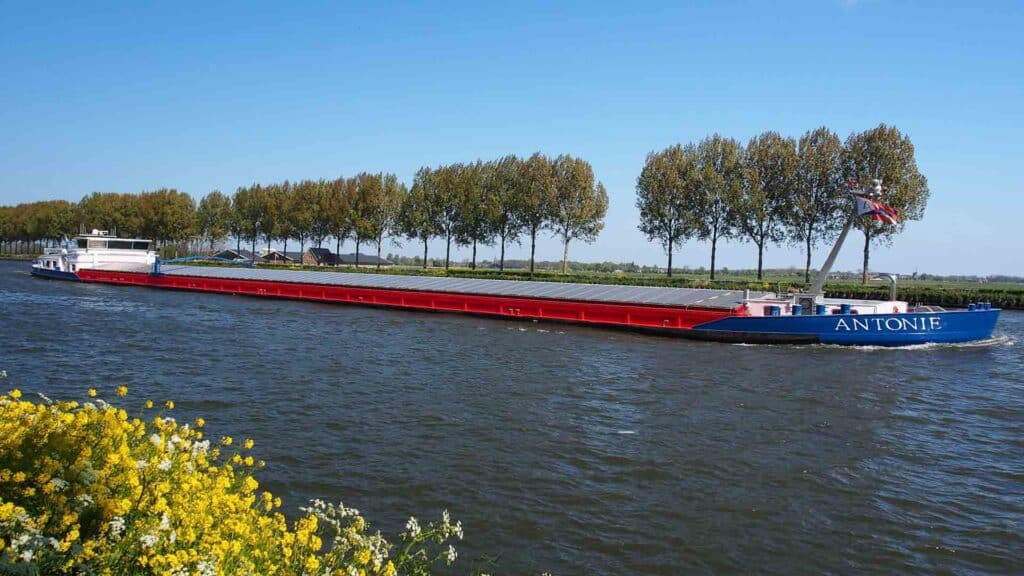 A longa barcaça de carga "ANTONIE" desliza pelo canal, ladeada por árvores e flores amarelas sob um céu azul claro — uma cena serena que pode refletir as tendências de transporte previstas para 2025.