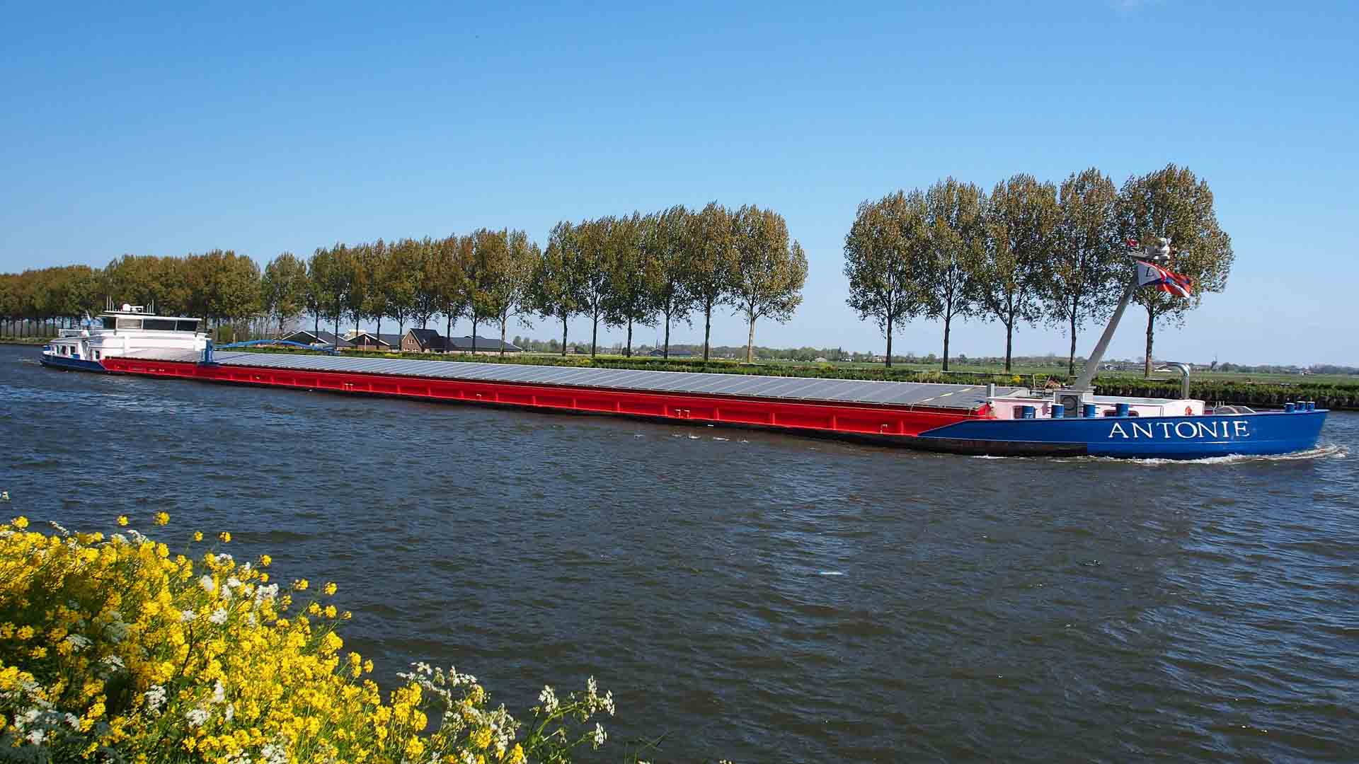 A longa barcaça de carga "ANTONIE" desliza pelo canal, ladeada por árvores e flores amarelas sob um céu azul claro — uma cena serena que pode refletir as tendências de transporte previstas para 2025.