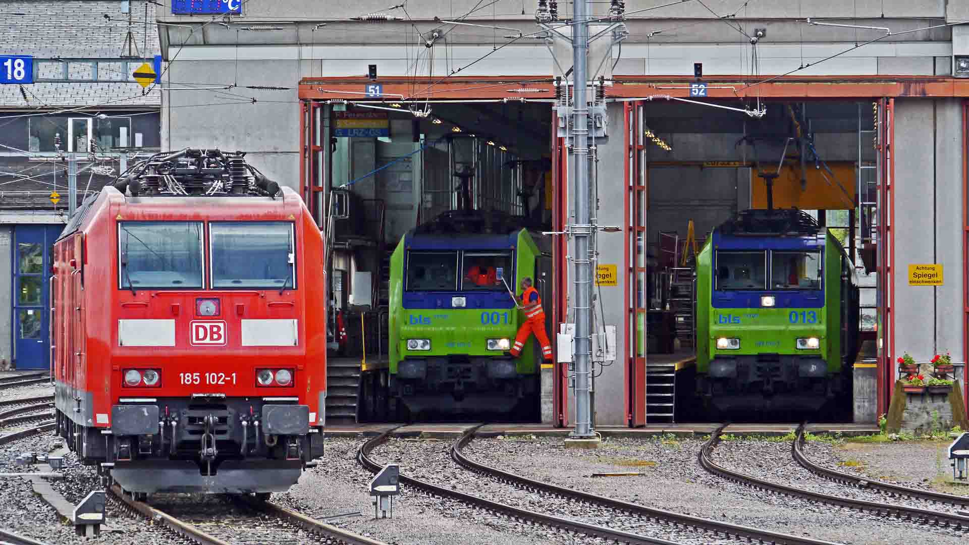 Um trem vermelho sobre trilhos fica perto de um prédio com três portas de garagem, onde dois trens verdes aguardam lá dentro. Uma pessoa está por perto, possivelmente supervisionando a importação de produtos lucrativos. Fios aéreos e sinalização completam essa cena movimentada.