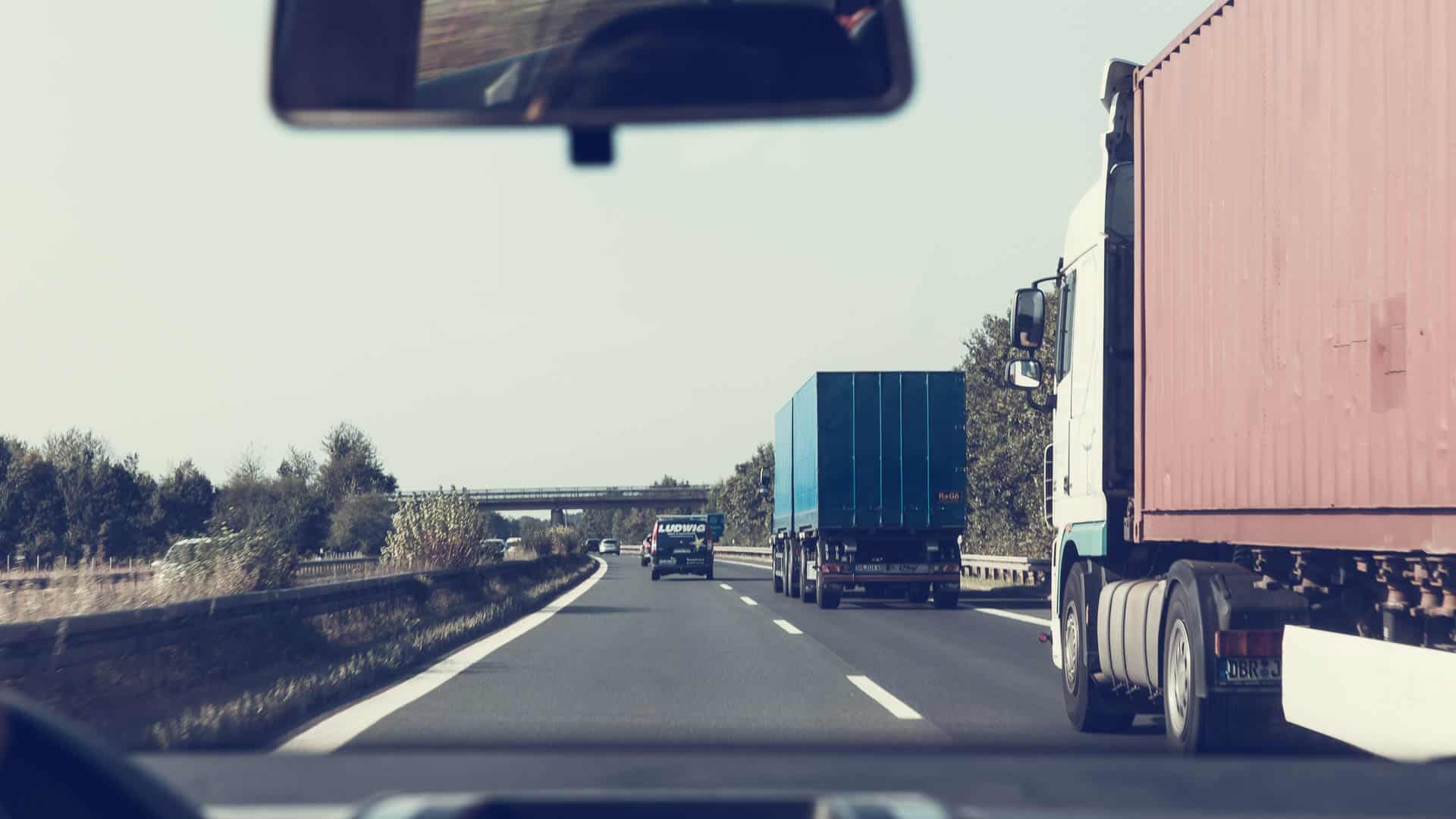 Vista do interior de um carro dirigindo em uma rodovia com dois caminhões grandes, possivelmente transportando produtos de alto crescimento, um em cada faixa. Um viaduto é visível à distância, simbolizando estradas futuras como aquelas que podemos pegar em 2025, à medida que as empresas continuam a importar mercadorias através das fronteiras.