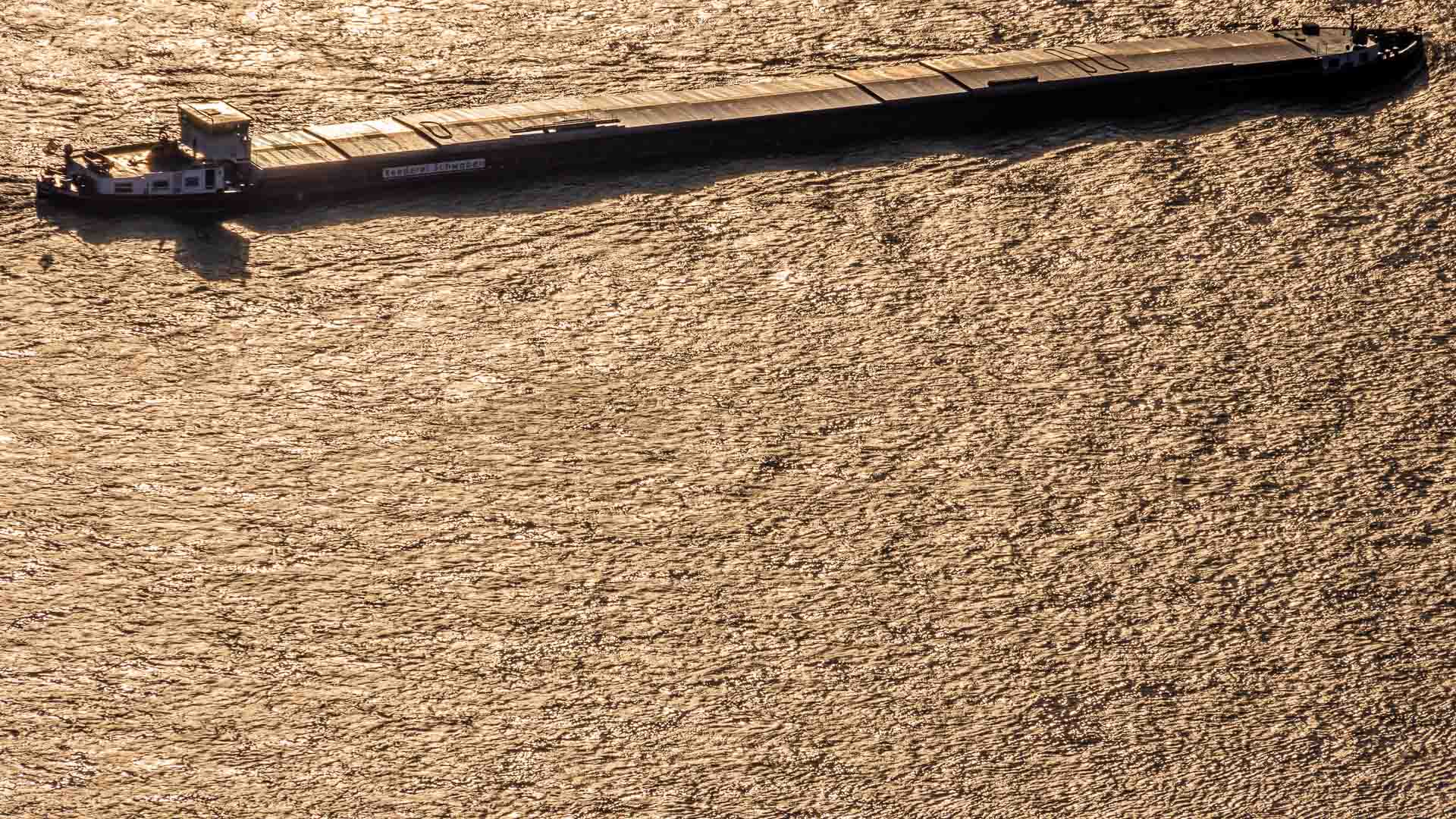 Vista aérea de um grande navio de carga, símbolo do comércio exterior, navegando por um corpo d'água iluminado pelo sol, criando suaves ondulações na superfície.