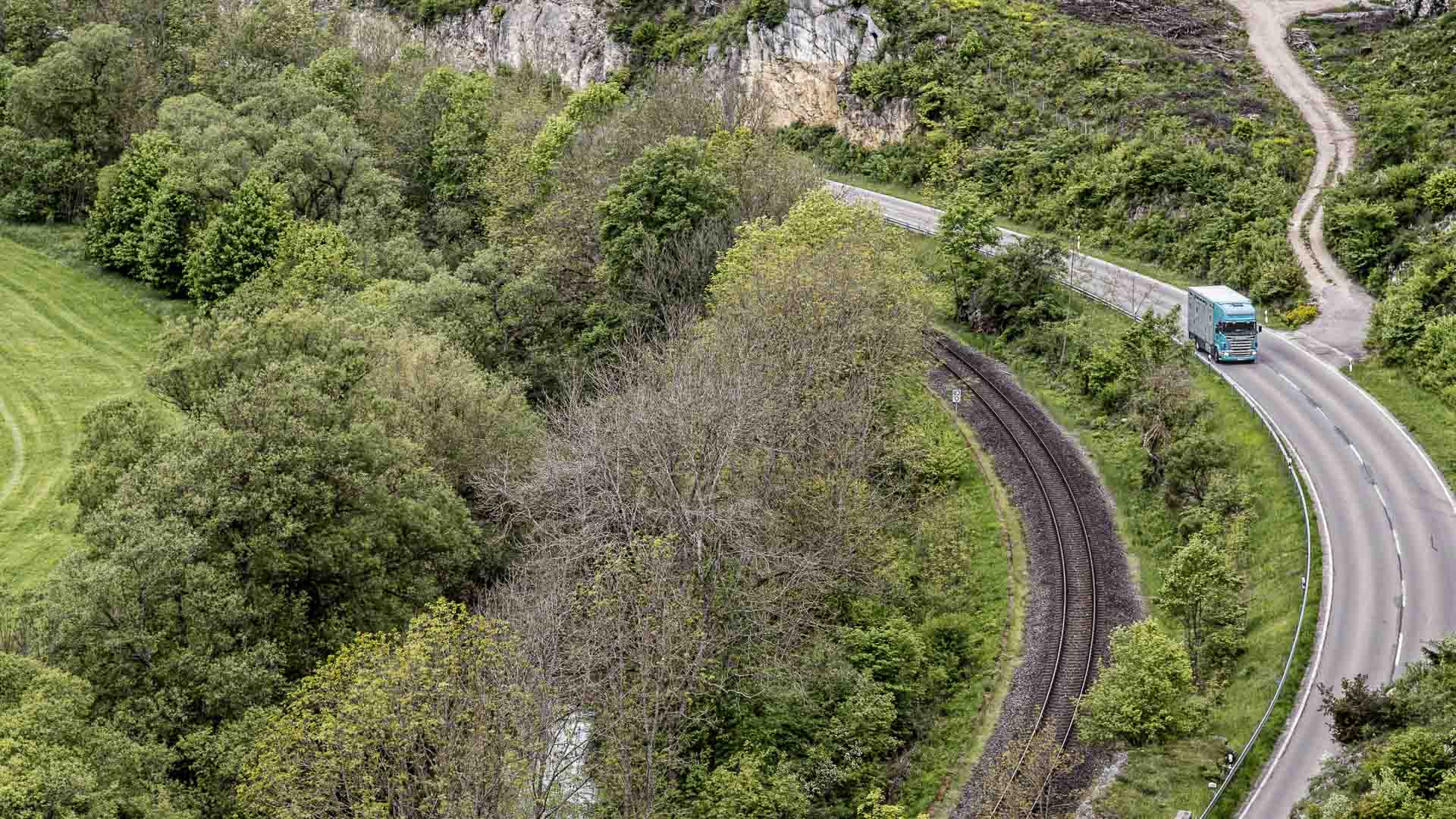 Vista aérea de uma estrada sinuosa e ferrovia cercada por vegetação densa e penhascos rochosos. Um caminhão azul, possivelmente transportando mercadorias certificadas pelo Inmetro, está viajando pela estrada.