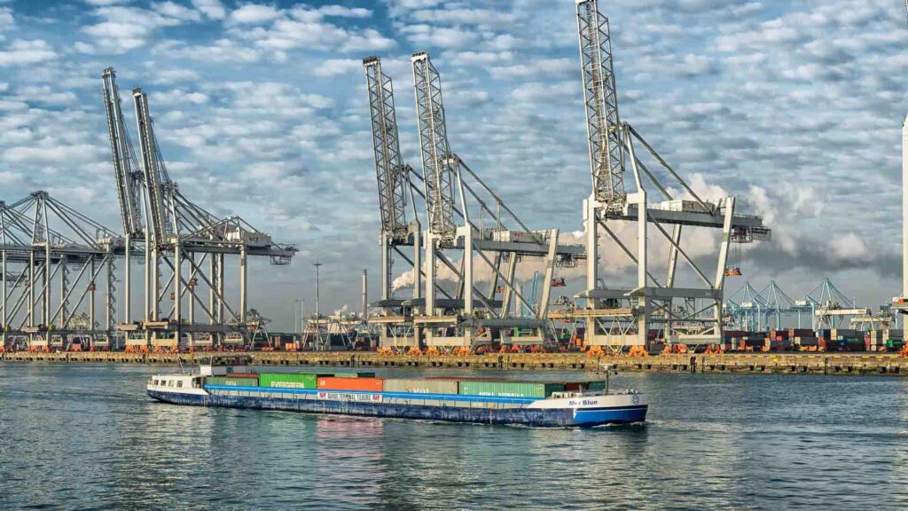 Em 2025, um navio de carga em um rio, carregado com contêineres coloridos, navega por grandes guindastes e estruturas industriais sob o céu nublado, mostrando a integração perfeita da tecnologia moderna na importação global.
