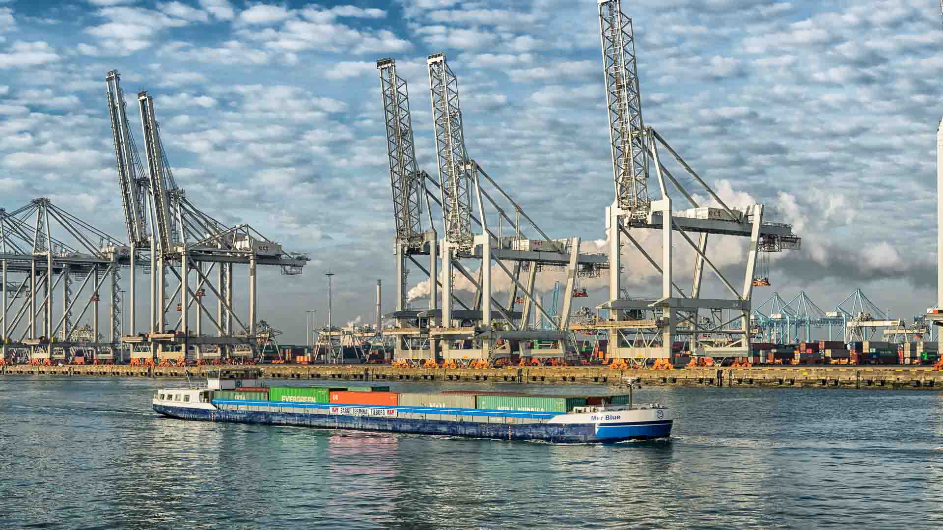 Em 2025, um navio de carga em um rio, carregado com contêineres coloridos, navega por grandes guindastes e estruturas industriais sob o céu nublado, mostrando a integração perfeita da tecnologia moderna na importação global.