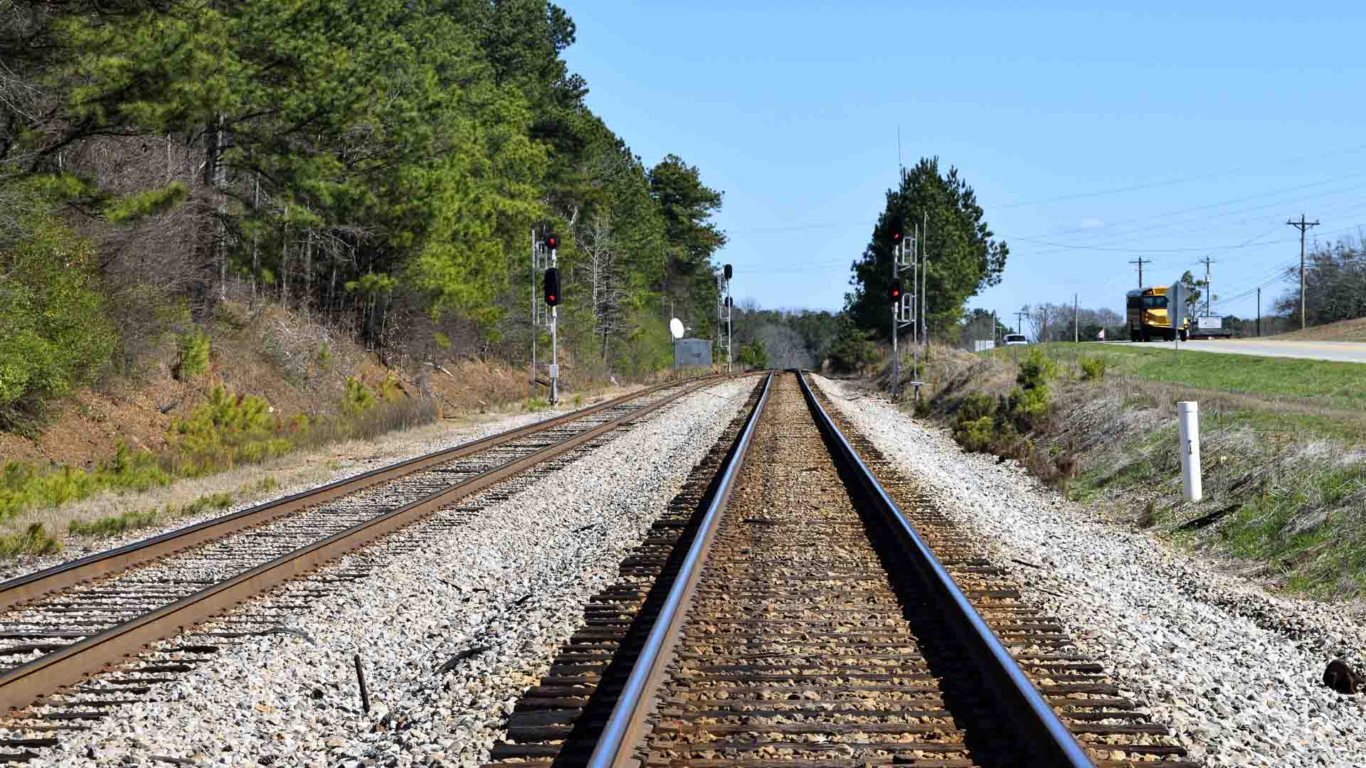 Os trilhos da ferrovia se estendem à distância, ladeados por árvores e grama de um lado, enquanto uma estrada com um veículo amarelo segue do outro sob o céu azul claro, incorporando o princípio do Tempo de Transporte nesta paisagem inspirada no Brasil e na China.